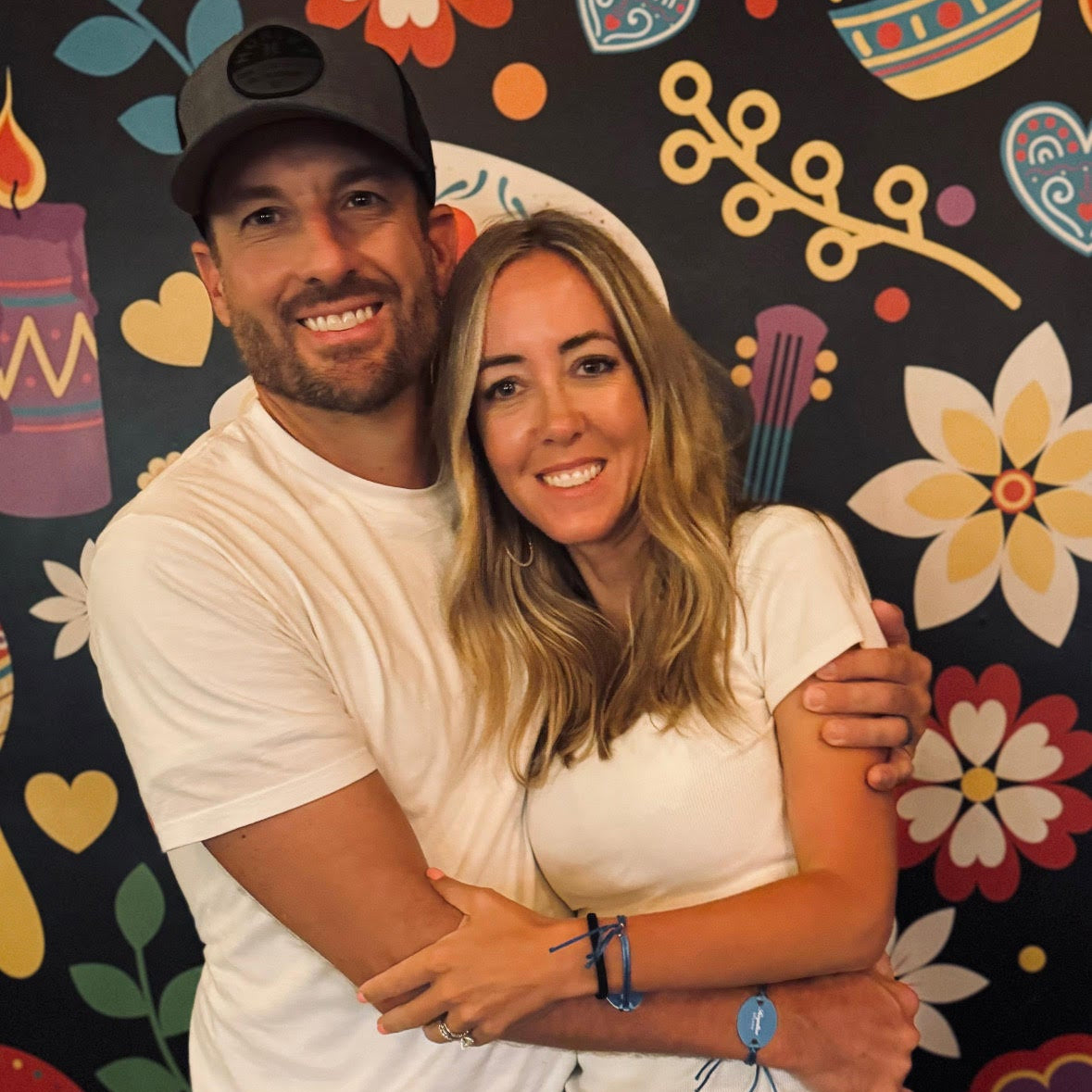 A smiling couple posing together in front of a colorful Day of the Dead themed wall mural. The man wears a black baseball cap, white t-shirt, and blue jeans, while the woman wears a white t-shirt and black pants.